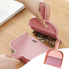 Pink mini hand brush and dustpan set cleaning debris from a desk surface for compact desktop cleanup.