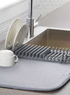 Dish Drying Mat with Rack, a super absorbent grey kitchen draining pad holding plates, cups, and silverware by a sink.