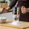 Stainless Steel Flour Sifter Cup for baking, handheld manual flour filter sifting flour onto a wooden cutting board.